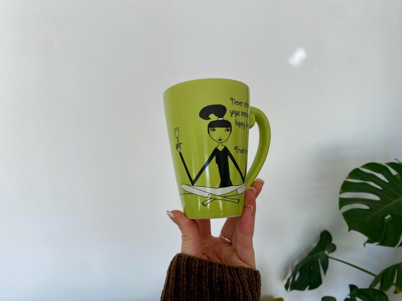 May include: A light green ceramic mug with a black handle. The mug features a cartoon illustration of a woman in a yoga pose holding a wine glass, with the text "Never skip yoga, happy hour, or Friday".