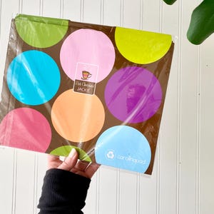 May include: A rectangular notebook with a brown background featuring large, colorful polka dots in shades of lime green, pink, yellow, turquoise, orange, and purple. A label in the center reads Hot Chocolate JACKIE and the bottom right says carolinapad.