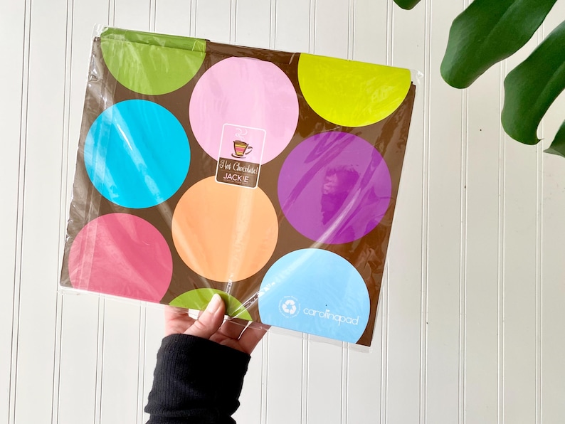 May include: A rectangular notebook with a brown background featuring large, colorful polka dots in shades of lime green, pink, yellow, turquoise, orange, and purple. A label in the center reads Hot Chocolate JACKIE and the bottom right says carolinapad.