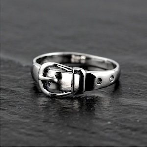May include: A silver ring with a buckle design. The buckle is detailed with a raised edge and a small, round, clear stone.