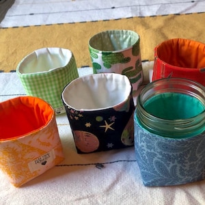 Op de afbeelding: Een verzameling stoffen potjeshoezen in verschillende patronen en kleuren. Ontwerpen omvatten een groene cactusprint, een zwarte ruimtescène, een paisley-print en een ruitpatroon. Elke hoes heeft een contrasterende binnenvoering en een label met "HALZEY".