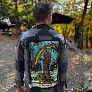 May include: A grey and black buffalo plaid flannel shirt featuring a large rectangular back patch. The patch displays a graphic of a character in a striped sweater and hat with clawed hands, a rainbow, houses, and the text Elm St and The Dreamer.