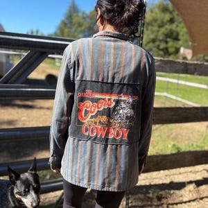 May include: A striped button-down shirt with a patch featuring "Coors Cowboy" text and graphic. The shirt has vertical stripes in shades of blue, red, and gray. The patch is black with red and white lettering.