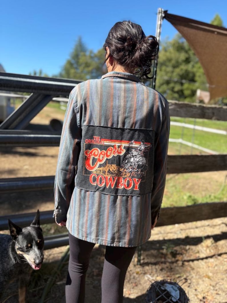 May include: A striped button-down shirt with a patch featuring "Coors Cowboy" text and graphic. The shirt has vertical stripes in shades of blue, red, and gray. The patch is black with red and white lettering.