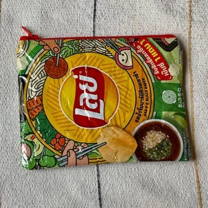May include: A rectangular zippered pouch made from a repurposed Lay's potato chip bag. The design features the Lay's logo, food illustrations, and text in a foreign language. The zipper is red.