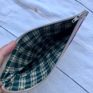 May include: Open beige zippered pouch with a green and tan plaid interior. The exterior has a colorful striped design on the side. The zipper is silver. The pouch is open, revealing the interior lining.
