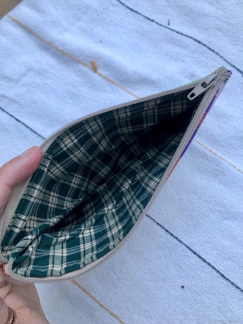 May include: Open beige zippered pouch with a green and tan plaid interior. The exterior has a colorful striped design on the side. The zipper is silver. The pouch is open, revealing the interior lining.