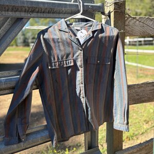 May include: A button-down shirt with vertical stripes in shades of blue, brown, and red hangs on a metal gate. The shirt has two chest pockets and long sleeves. A white tag with the word "Thank" is attached to the shirt.