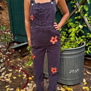 May include: Dark purple denim overalls featuring a straight-leg silhouette, a front bib pocket, and metal hardware on the shoulder straps. The garment is decorated with multiple orange and pink fabric flower appliques scattered across the front.
