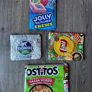 May include: Four small zippered pouches made from repurposed food packaging. The pouches feature the logos and designs of Jolly Rancher Chews, Klondike Mint Chocolate Chip, Lay's, and Tostitos Salsa Verde. The pouches are rectangular and have a zipper closure.