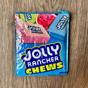 May include: A rectangular, blue zippered pouch featuring a Jolly Rancher Chews design. The pouch displays images of watermelon, pink, and blue Jolly Rancher candies. The text "JOLLY RANCHER CHEWS" is prominently displayed in white and yellow.