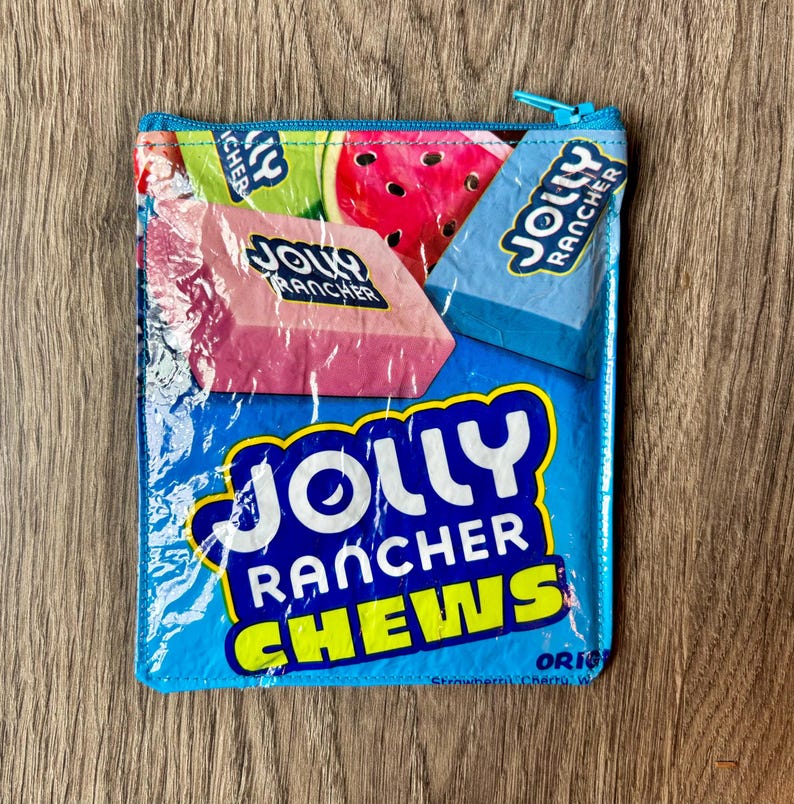 May include: A rectangular, blue zippered pouch featuring a Jolly Rancher Chews design. The pouch displays images of watermelon, pink, and blue Jolly Rancher candies. The text "JOLLY RANCHER CHEWS" is prominently displayed in white and yellow.