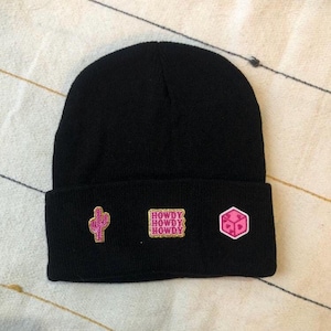 May include: Black beanie hat with three embroidered patches. The patches include a pink cactus, the word "HOWDY" repeated three times, and a pink geometric shape with hearts. The hat is made of a soft, knitted material.