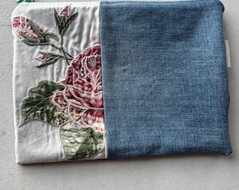 May include: A rectangular zippered pouch featuring a split design. The left side is white fabric with an embroidered pink rose and green leaves. The right side is blue denim fabric. A small mint green flower-shaped zipper pull is attached.