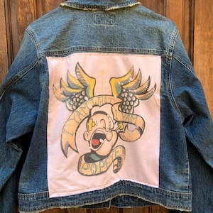 May include: A denim jacket with a light blue wash. The back features a large patch with a Popeye the Sailor graphic, including the text "I Yam What I Yam" and "Popeye". The graphic is in shades of yellow, green, and black on a pink background.