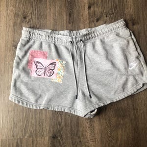 May include: Gray Nike shorts with a drawstring waist. A butterfly patch is sewn on the front, featuring a black butterfly design on a pink and floral background. The Nike logo is visible on the right leg.