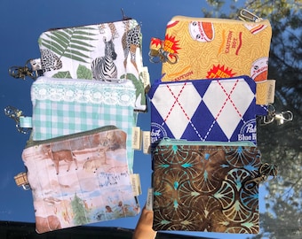 May include: Six small fabric zipper pouches with metal clips and Holzey tags. Designs include a zebra and giraffe print, a yellow ramen noodle pattern, a light blue gingham with lace, a blue argyle, a deer forest scene, and a dark fan pattern.
