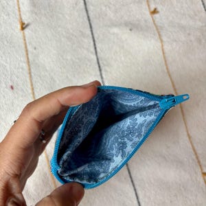 May include: Open, turquoise-zippered pouch with a blue floral patterned interior. The pouch is held open, revealing the inside. The background is a cream-colored textile with gold and black stripes.