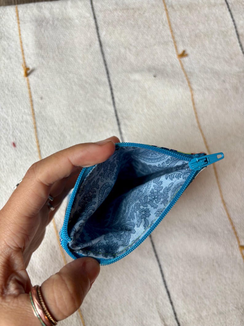 May include: Open, turquoise-zippered pouch with a blue floral patterned interior. The pouch is held open, revealing the inside. The background is a cream-colored textile with gold and black stripes.