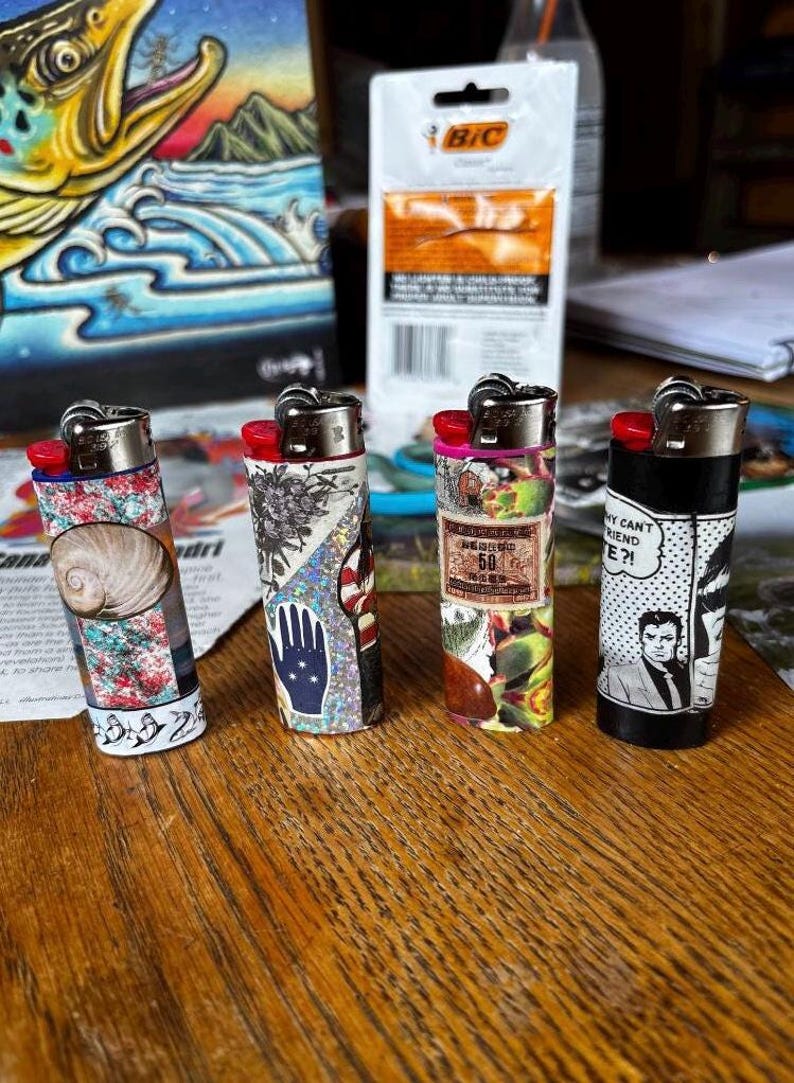 May include: Four decorative lighters with unique designs. One features a seashell, another a hand with a star pattern, a third with a vintage label, and the last with a comic strip design. Each lighter has a red ignition button.