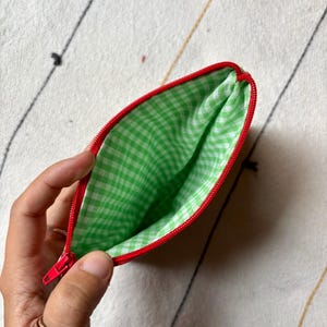 May include: A small, open pouch with a red zipper and trim. The interior is lined with green and white gingham fabric. The pouch is held open, revealing the inside. The background is a cream-colored fabric with black and gold lines.