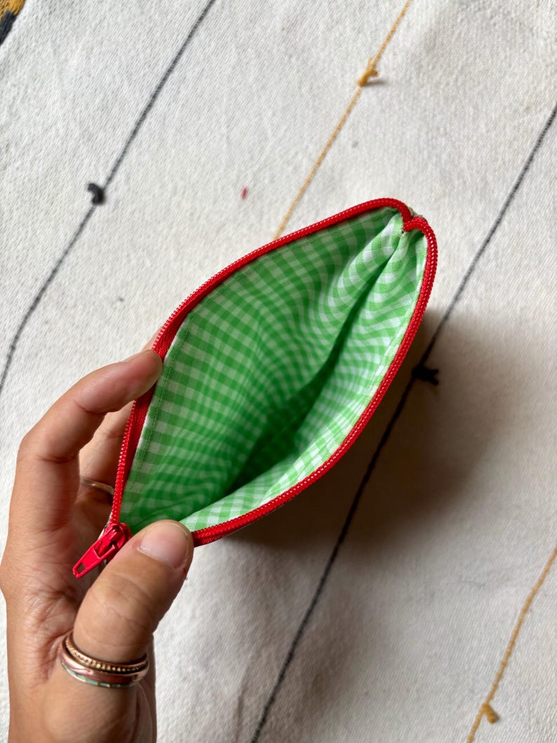May include: A small, open pouch with a red zipper and trim. The interior is lined with green and white gingham fabric. The pouch is held open, revealing the inside. The background is a cream-colored fabric with black and gold lines.
