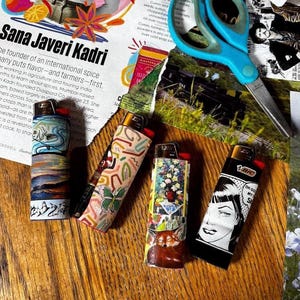 May include: Four BIC lighters with unique designs are displayed on a wooden surface. One features a pop art woman, another a floral arrangement, and the others have abstract patterns. A pair of blue scissors and a magazine are in the background.