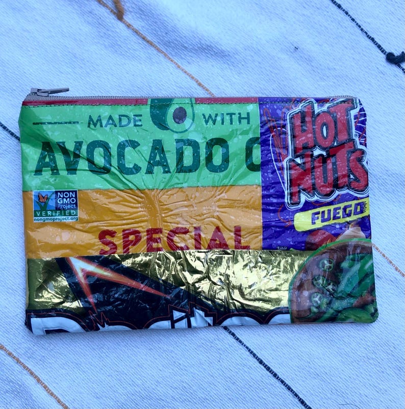 May include: A rectangular zippered pouch made from repurposed food packaging. The pouch features text that reads "MADE WITH AVOCADO OIL" and "HOT NUTS FUEGO". The pouch is primarily green, yellow, and purple.