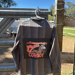 May include: A long-sleeved button-down shirt with vertical stripes in blue, gray, and orange. The back features a dark gray patch with the words "Coors Cowboy" and a graphic of a cowboy.
