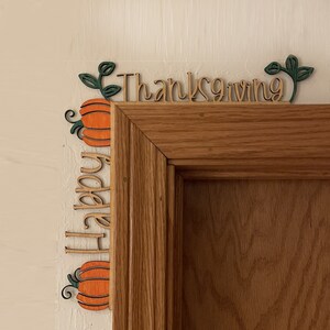 K&ouml;nnte beinhalten: Holz-T&uuml;rdekoration zum Erntedankfest mit den Worten "Happy Thanksgiving" und K&uuml;rbismotiven. Die K&uuml;rbisse sind orange mit gr&uuml;nen Bl&auml;ttern und schwarzen Ranken. Der Text ist in Schreibschrift.