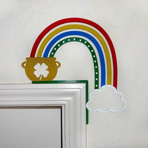 May include: A wooden rainbow with red, yellow, blue, and green stripes, with a gold pot of gold at the end. The rainbow is attached to a white door frame.