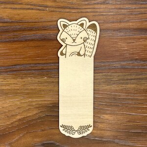 K&ouml;nnte beinhalten: Ein Lesezeichen aus Holz mit einer Laserschnitt-Illustration eines Waschb&auml;ren. Der Waschb&auml;r befindet sich oben auf dem Lesezeichen, darunter ein leerer rechteckiger Bereich f&uuml;r Notizen. Unten befindet sich ein dekoratives Blattdesign.