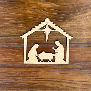 May include: A wooden cutout of a nativity scene with a star above the stable. The figures of Mary, Joseph, and the baby Jesus are visible inside the stable.