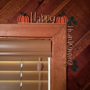 K&ouml;nnte beinhalten: Holz-Thanksgiving-Dekoration mit den Worten "Happy Thanksgiving". Die Dekoration zeigt orangefarbene K&uuml;rbisse mit gr&uuml;nen Stielen und Bl&auml;ttern. Die Dekoration befindet sich &uuml;ber einem Fensterrahmen.