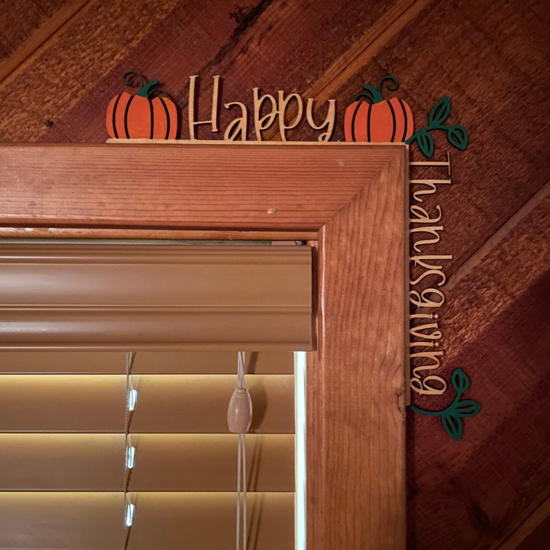 K&ouml;nnte beinhalten: Holz-Thanksgiving-Dekoration mit den Worten "Happy Thanksgiving". Die Dekoration zeigt orangefarbene K&uuml;rbisse mit gr&uuml;nen Stielen und Bl&auml;ttern. Die Dekoration befindet sich &uuml;ber einem Fensterrahmen.