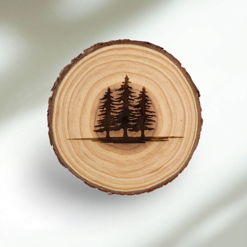 May include: A wooden coaster with a burnt design of three pine trees on a natural wood background.