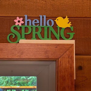 May include: A wooden sign with the words "hello spring" in green, blue, and pink letters. A yellow bird with a black eye is on the right side of the sign. The sign is attached to a wooden door frame.