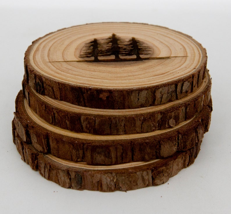 May include: Four wooden coasters stacked on top of each other. The top coaster has a woodburned design of three pine trees.