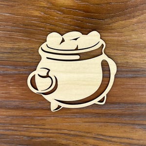 May include: A wooden cutout of a pot filled with gold coins. The pot has a handle and a rounded bottom. The cutout is a light brown color.