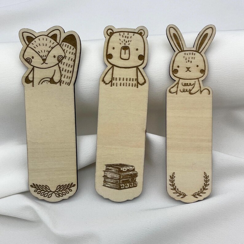K&ouml;nnte beinhalten: Drei Lesezeichen aus Holz mit Tierdesigns. Ein Fuchs, ein B&auml;r und ein Kaninchen sind abgebildet. Jedes Lesezeichen ist hellbraun mit einem schwarzen Rand. Dekorative Elemente sind enthalten.
