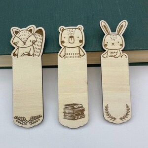 K&ouml;nnte beinhalten: Drei Lesezeichen aus Holz mit Tierdesigns: ein Fuchs, ein B&auml;r und ein Kaninchen. Der Fuchs hat einen buschigen Schwanz, der B&auml;r hat ein gen&auml;htes Muster und das Kaninchen hat lange Ohren. Jedes Lesezeichen hat einen leeren rechteckigen Bereich.