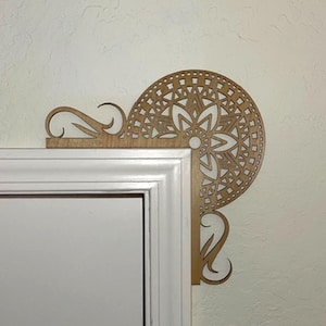 May include: A wooden corner accent piece with a floral mandala design. The decorative piece is light brown and features intricate cut-out patterns. It is mounted on a white door frame and a light beige wall.