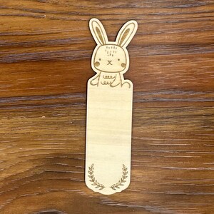 K&ouml;nnte beinhalten: Ein Lesezeichen aus Holz mit einem lasergeschnittenen Design eines Cartoon-Hasen. Der Hase hat detaillierte Merkmale. Der untere Teil des Lesezeichens hat ein Lorbeerkranz-Design. Das Lesezeichen ist aus hellem Holz gefertigt. Ca. 15 cm lang.