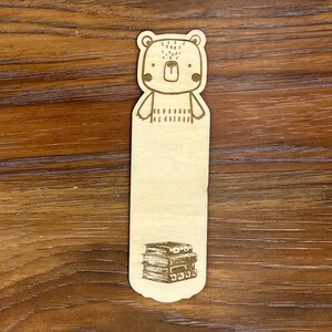 K&ouml;nnte beinhalten: Ein Lesezeichen aus Holz mit einer B&auml;renillustration oben und einem B&uuml;cherstapel unten. Der B&auml;r hat ein einfaches Design mit gepunkteten Details und rosigen Wangen. Das Lesezeichen ist aus hellem Holz gefertigt.