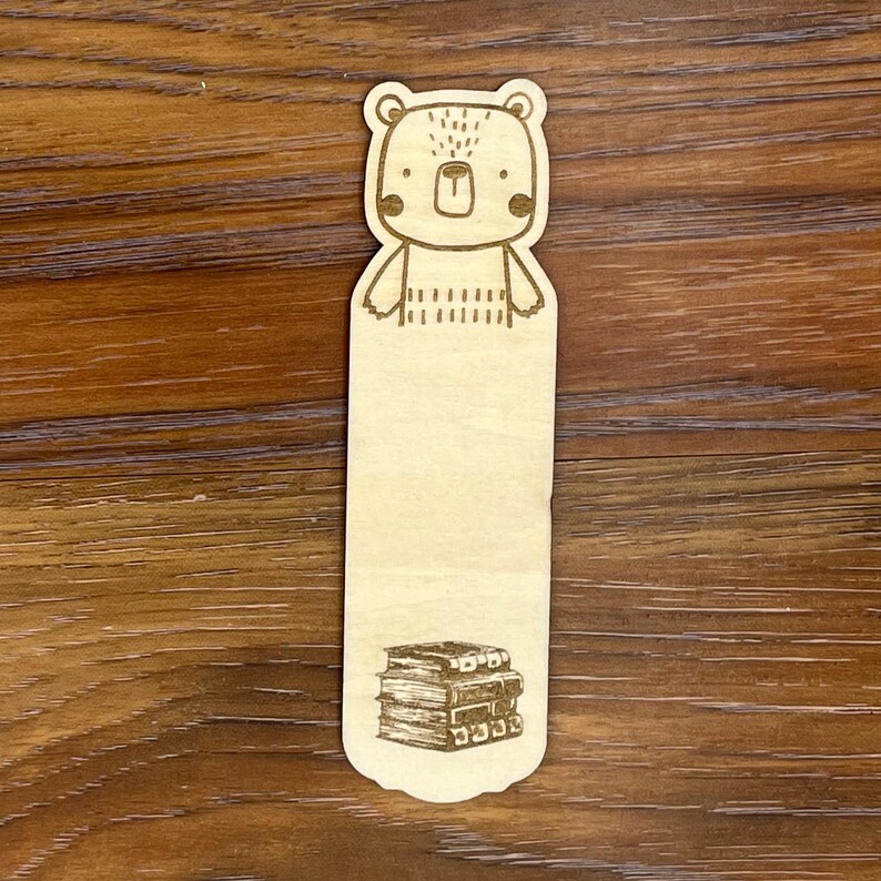 K&ouml;nnte beinhalten: Ein Lesezeichen aus Holz mit einer B&auml;renillustration oben und einem B&uuml;cherstapel unten. Der B&auml;r hat ein einfaches Design mit gepunkteten Details und rosigen Wangen. Das Lesezeichen ist aus hellem Holz gefertigt.