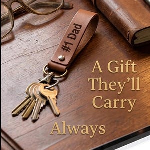 May include: A brown leather keychain with the text "#1 Dad" and a set of keys. The image also includes a notebook, glasses, and the text "A Gift They'll Carry Always."