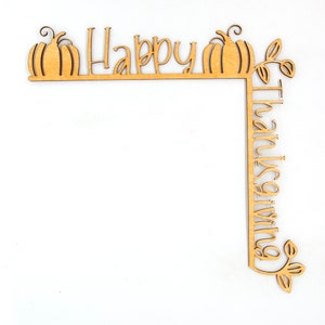 K&ouml;nnte beinhalten: Eckdekoration aus Holz mit den Worten "Happy Thanksgiving" ausgeschnitten. Das Design umfasst zwei K&uuml;rbisse und Bl&auml;tterakzente. Das Holzst&uuml;ck ist hellbraun und liegt auf einem wei&szlig;en Hintergrund.