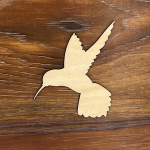 May include: A wooden cutout of a hummingbird in flight. The cutout is a light brown color and has a detailed shape.