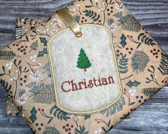 May include: A gift tag with the name "Christian" embroidered in brown thread, featuring a green Christmas tree design. The tag is attached to a wrapped gift with a gold ribbon, placed on a brown paper with a botanical pattern.
