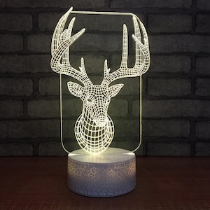 Puede incluir: Una luz nocturna de cabeza de ciervo 3D blanca con una base blanca. La cabeza de ciervo está hecha de acrílico transparente y tiene un diseño de estructura de alambre. La base tiene una superficie texturizada.